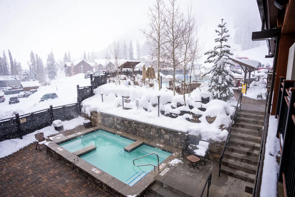 An inviting outdoor hot tub set amidst a snowy landscape and pine trees. The stone steps and serene surroundings evoke relaxation and togetherness, especially after winter adventures. The hot tub is a social hub for families to unwind and bond.