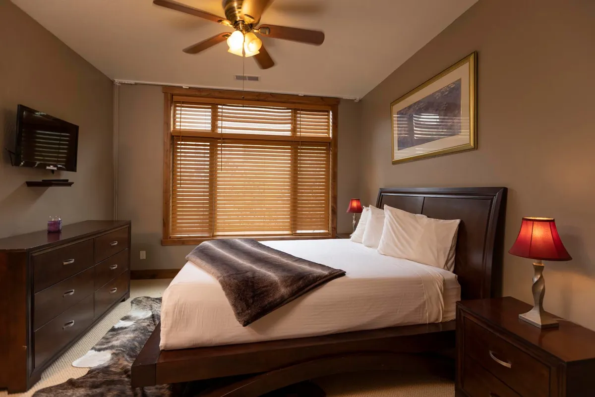 A warmly lit bedroom featuring a plush, inviting bed with layered blankets and pillows, complemented by wooden furniture and bedside lamps with striking red shades. A window with blinds allows natural light to filter in, highlighting the room's cozy, mountain-inspired details.
