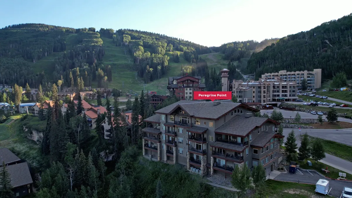 A wide-angle exterior shot of the Peregrine Point building nestled among tall evergreen trees with a dramatic mountain backdrop, lightly dusted with snow. The building stands just steps away from the ski slopes, capturing the unbeatable slopeside location.
