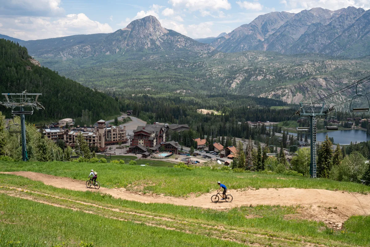 A panoramic exterior scene featuring a dramatic mountain range, a bike trail winding past the resort buildings, a tranquil lake, and a visible ski lift. The resort's proximity to nature and adventure activities is clearly depicted, with lush greenery and outdoor spaces ideal for family exploration and fun.