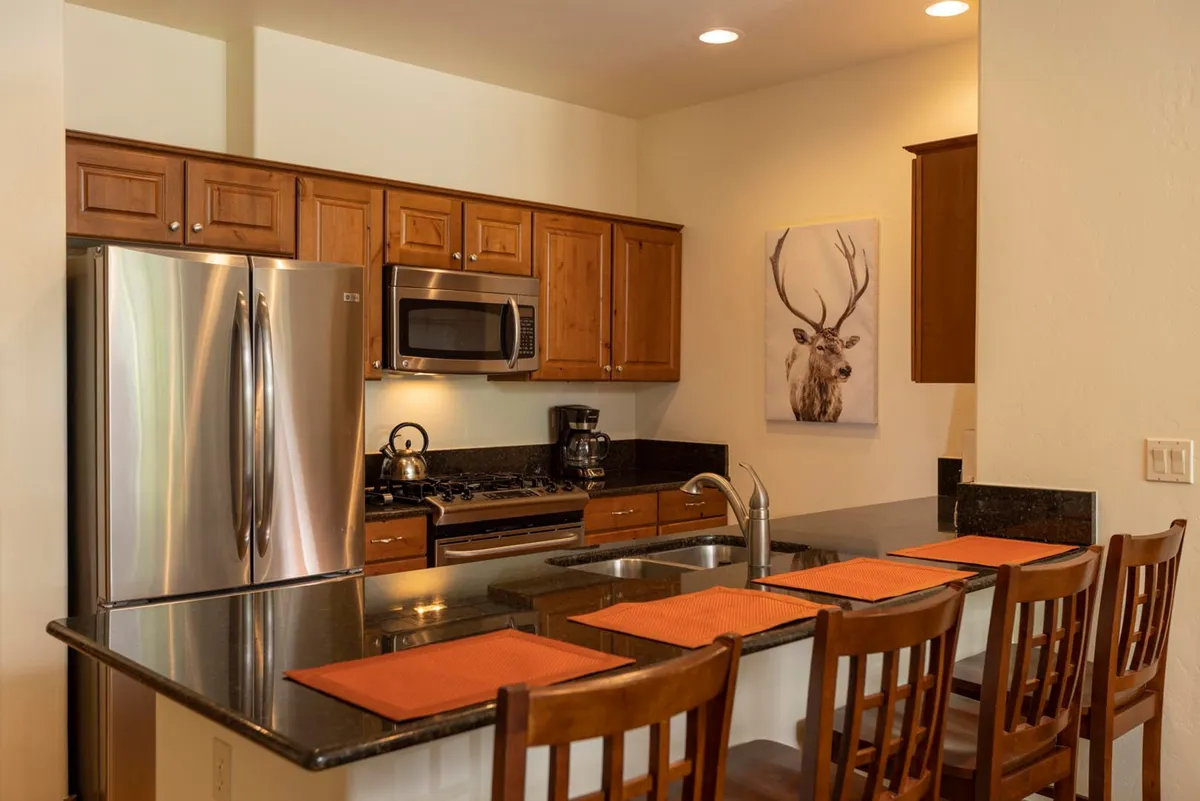 A spacious kitchen area featuring a central island topped with granite countertops and surrounded by modern stainless steel appliances. The scene includes tasteful mountain-themed artwork (notably a deer), with elements set up to evoke activities like cooking, entertaining guests, and sharing family meals. Warm lighting and cozy decor contribute to a welcoming ambiance.