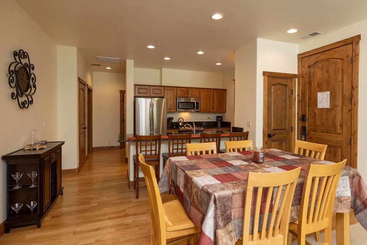 A warm and homey dining area featuring a dining table with a patterned tablecloth, surrounded by wooden cabinetry and stainless steel appliances. A decorative wall clock adds a touch of personality, and the space is inviting, ideal for family meals and social gatherings.