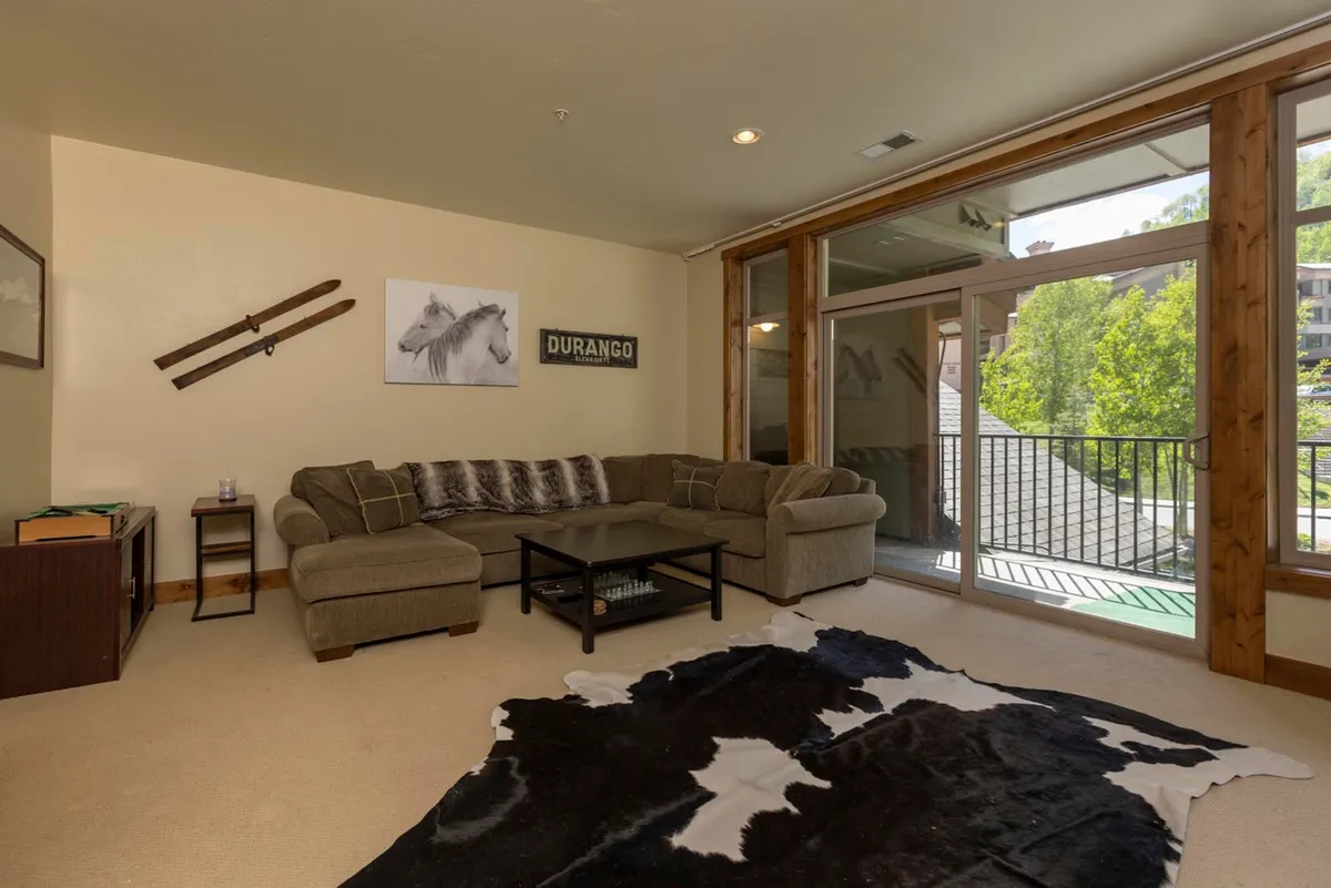 A spacious living room with plush sofas, a coffee table atop a cowhide rug, and ski-themed decor. Large windows flood the space with natural light and offer views of the surrounding landscape. The ambiance is welcoming, with cozy furnishings and a visible fireplace that anchors the area as the heart of the home.