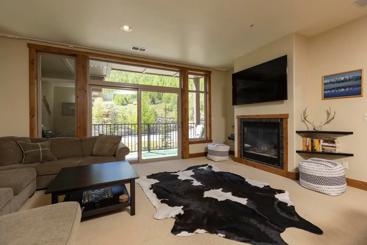 The image showcases a luxurious condo living room with a plush sofa, a glowing fireplace, a stylish cowhide rug, and expansive windows framing breathtaking mountain views. Soft natural light fills the space, creating a warm, inviting atmosphere perfect for relaxation. The balcony promises awe-inspiring sunsets and serene moments with a hot drink, all just steps from the ski lifts and resort plaza.
