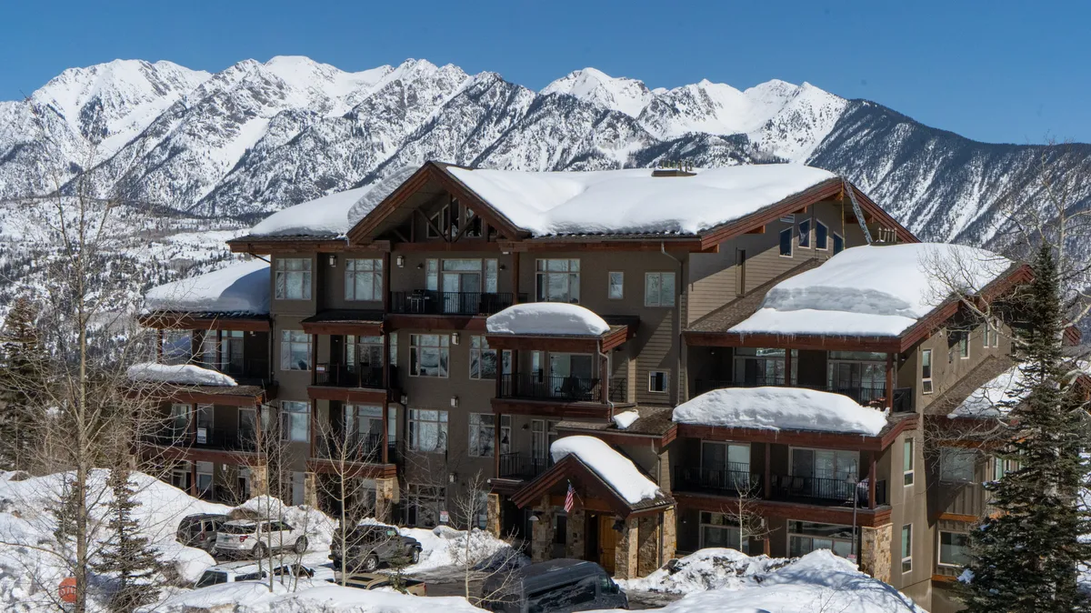 An exterior view featuring snow-covered peaks, attractive building architecture, and surrounding trees. The scene embodies the excitement of ski-in, ski-out convenience and the immediate proximity to adventure.