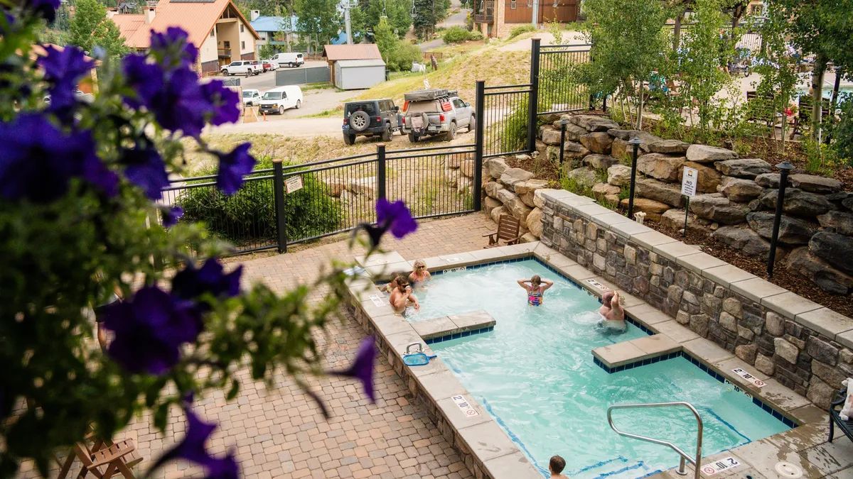 A vibrant exterior shot of a swimming pool surrounded by blooming flowers, with families and friends enjoying the water. The space is inviting, lively, and surrounded by nature, offering a communal atmosphere for relaxation and play.