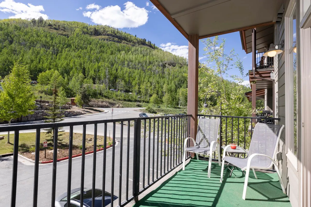 An outdoor balcony with a sturdy railing and comfortable chairs, overlooking a lush green landscape and distant mountains. The seating invites you to enjoy morning coffee, an evening toast, or simply soak in the peaceful mountain scenery.